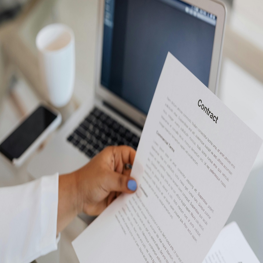 A woman's hand holding a contract. On the desk next to her is an open laptop, a mobile phone and a mug.