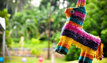 A colorful piñata shaped like a donkey hanging outdoors with a lush green background and a few blurred balloons in the distance.