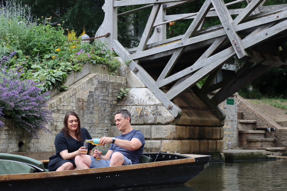 A couple on a punt pooring a cider into two glasses in front of the Mathematical Bridge on the river cam.