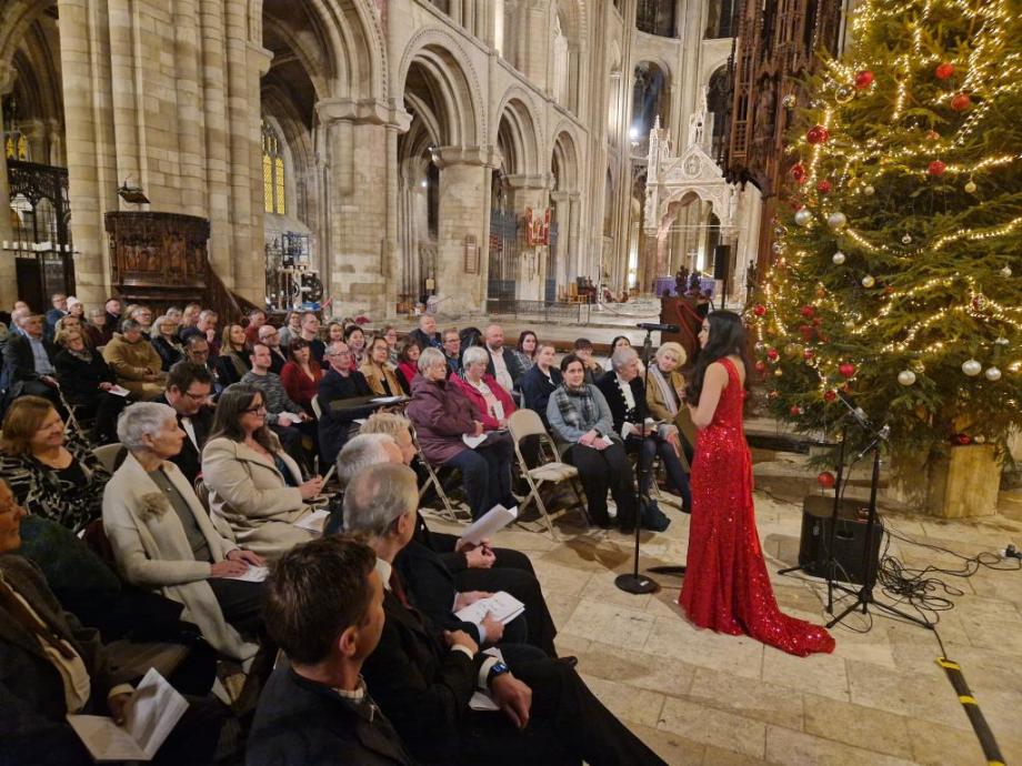 Singer, Gabriella Pineda-Rodrigues, in a red sequined dress performing in front of guests inside Peterborough Cathedral. A decorated Christmas tree is next to the singer.