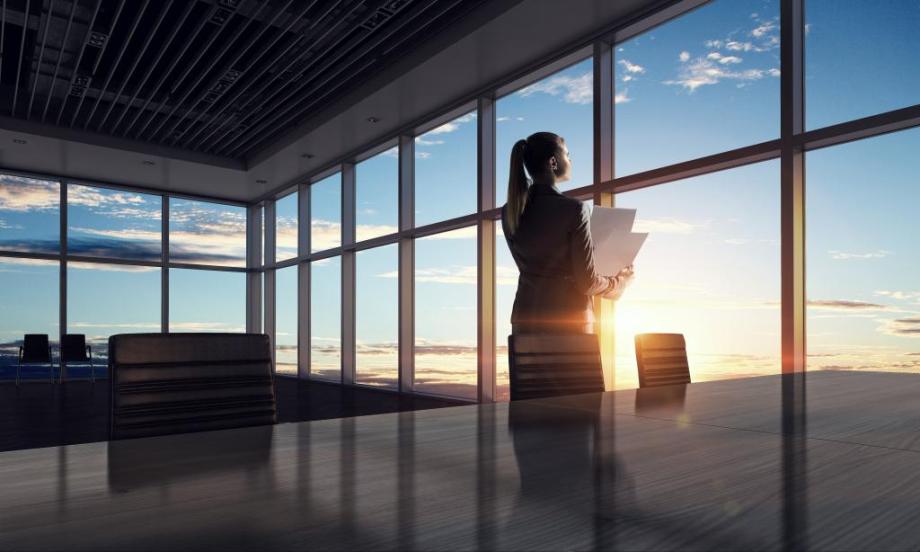 Women in corporate setting looking out office building at sunrise with paperwork in hand.