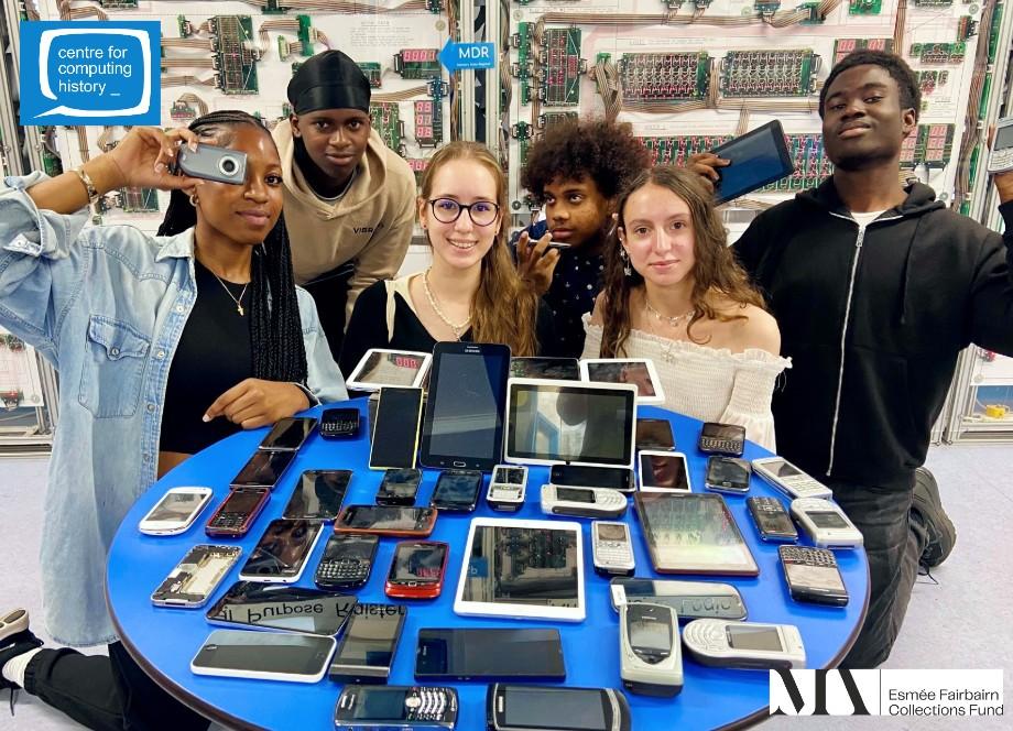 A group of young people at the Centre for Computing History, behind a table of old mobile phones and tablets ready for recycling