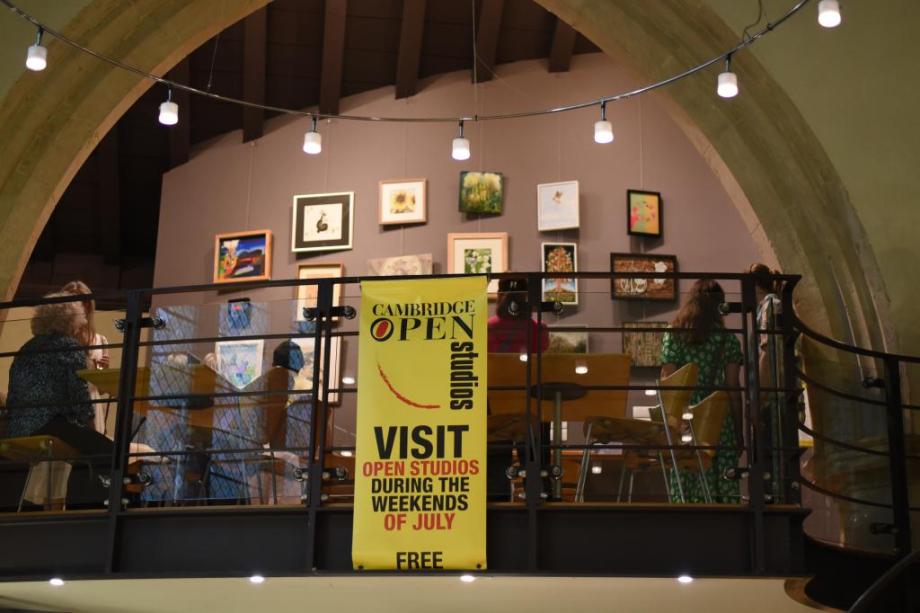 Image depicts Cambridge Open Studios yellow banner in front of display of paintings 