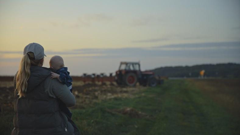 Mother holds baby while overlooking a tractor in field at sunset 