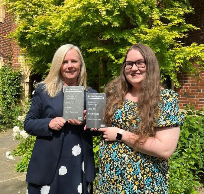 x2 white women standing in a lush green garden holding a grey slate award for Green Impact