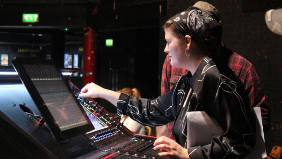 A young woman wearing a black shirt is at a mixing desk, adjusting the controls.