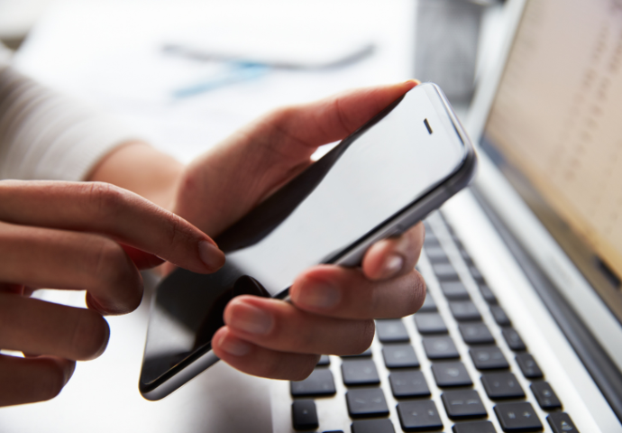 hand holding a mobile phone with a laptop in the background