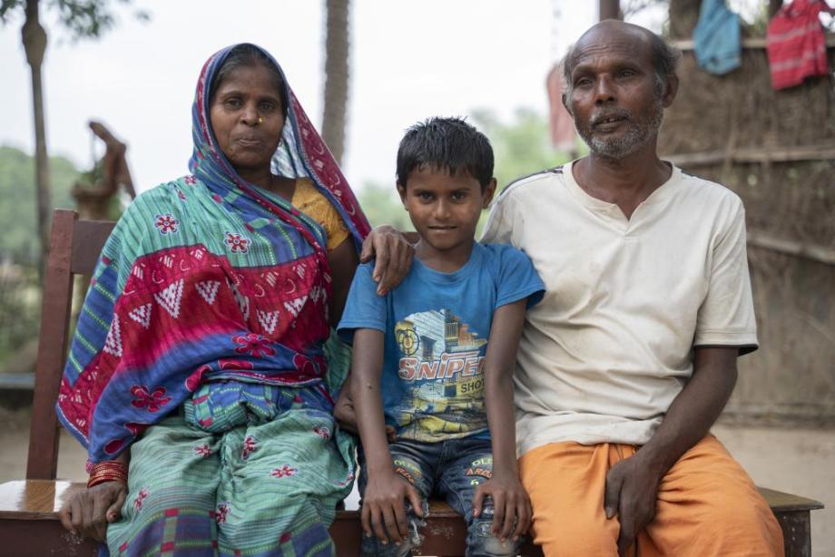 Aanand, wearing a blue t-shirt sits between his parents outside. His mother is resting her hand on his shoulder.