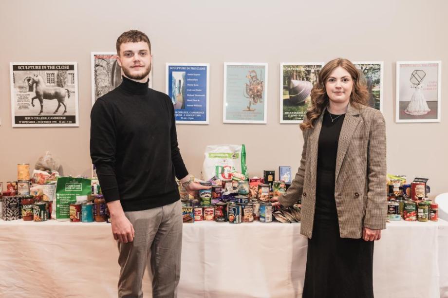 Ryan Gooda & Sophie Edwards from meet Cambridge standing in front of a long white table clothed table with food donations