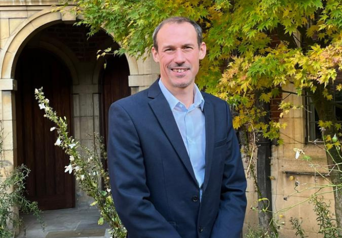 White man in a dark blue suit smiling at the camera outside in front of an old Cambridge College with greenery in the background. Andrew Bell the new Head of Meet Cambridge