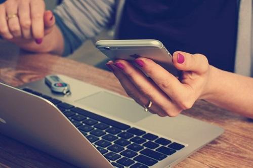 woman's hand holding mobile phone and hovering over computer keyboard