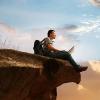 Man with laptop sitting on cliff edge and staring at the sky